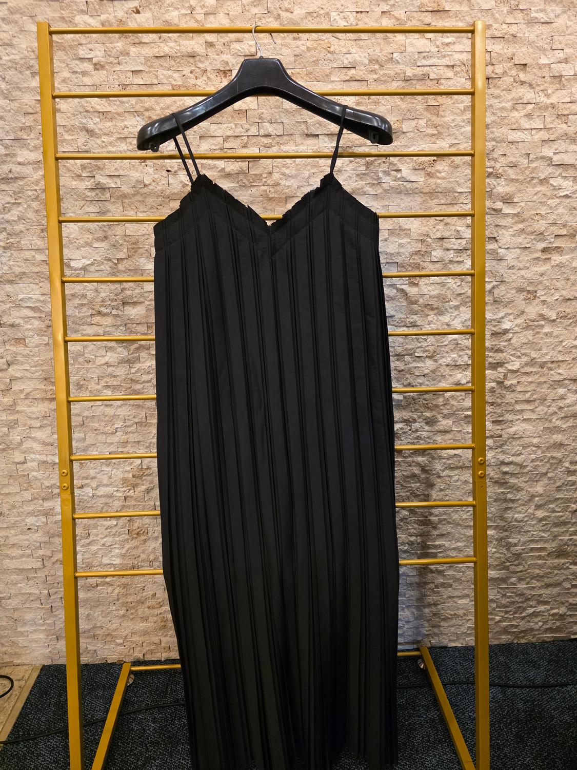 Pleated black dress hanging on a black hanger on a gold structure, with a light brick wall background.
