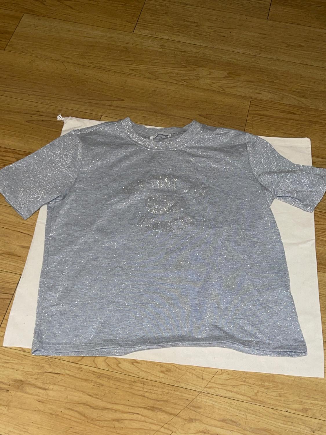 Gray glitter t-shirt with an eye design in the center, placed on a light wooden surface.