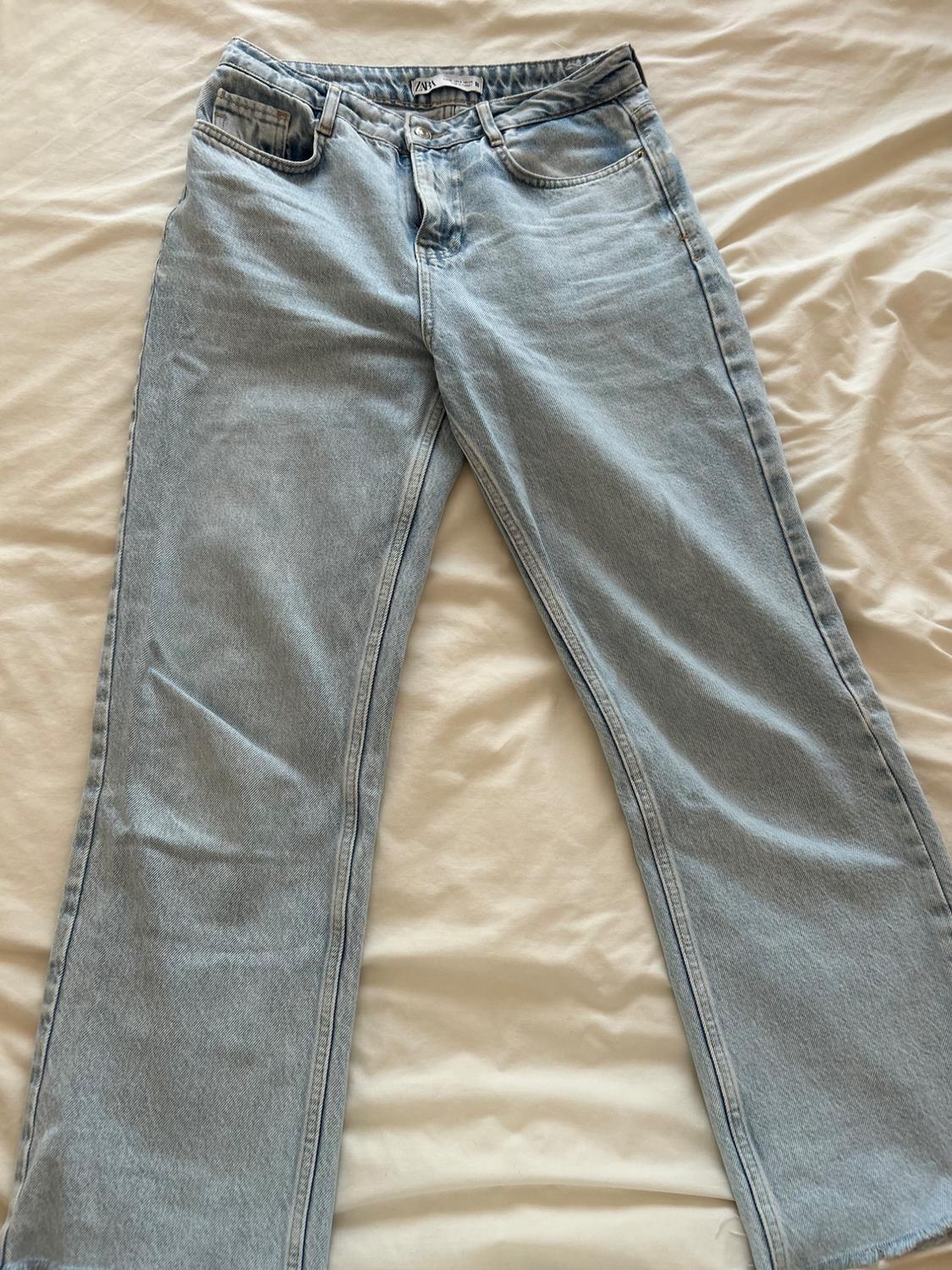 Light blue denim jeans with a chequered front pocket, placed on a beige surface. Visible tag on the waistband.