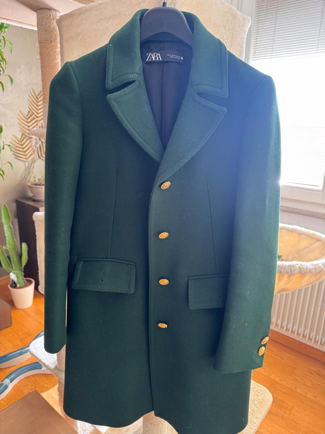 Dark green coat with gold buttons hanging on a hanger. Background with plants and natural light coming through the window.