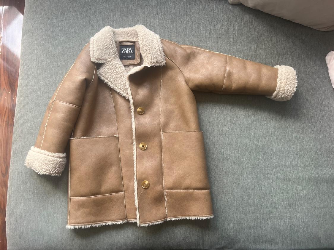 Light brown jacket with a white lining, collar and cuffs. Three buttons, pockets and a ZARA label. It rests on a grey surface.