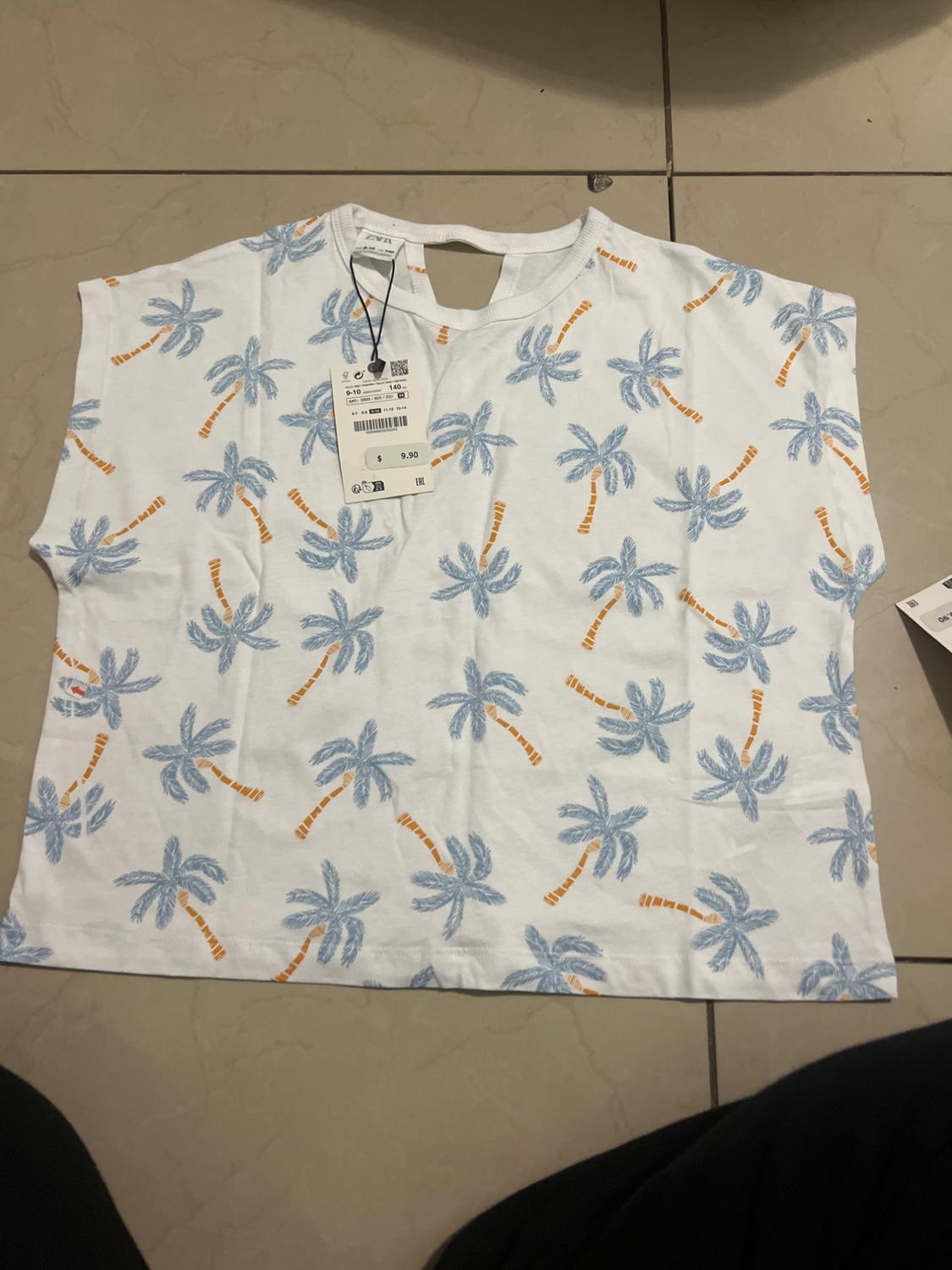 White T-shirt with blue palm tree print and orange trunks on a tiled floor. Price tag of 9.90 euros visible.