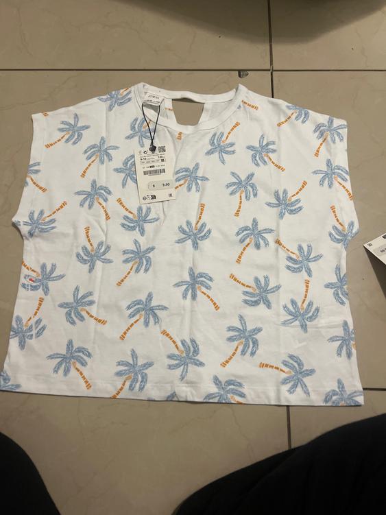 White t-shirt with blue palm tree print and orange trunks on a tiled floor. Price tag of 9.90 euros visible.