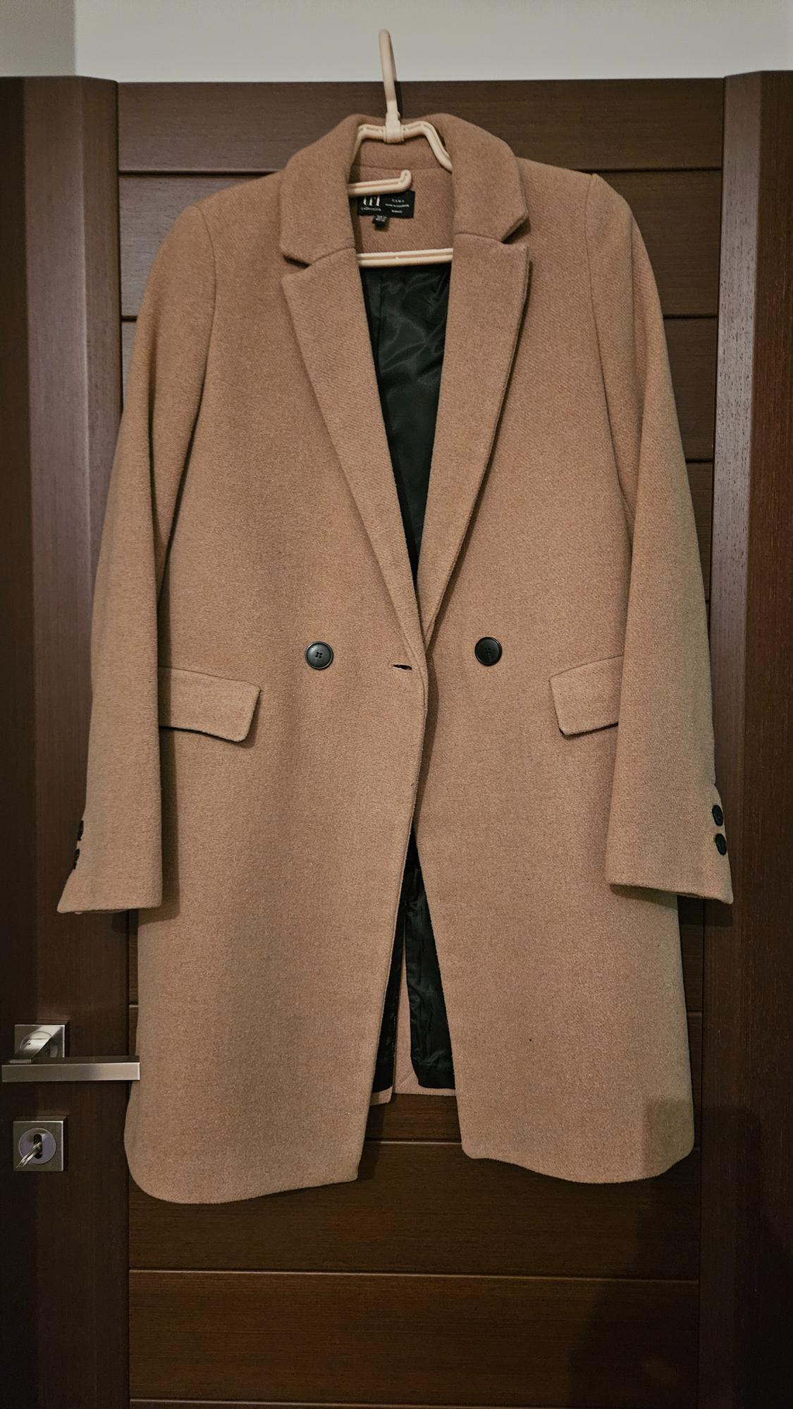 Camel coat hanging on a dark wooden door. It has lapels, two black buttons, and flap pockets. Black inner lining visible.
