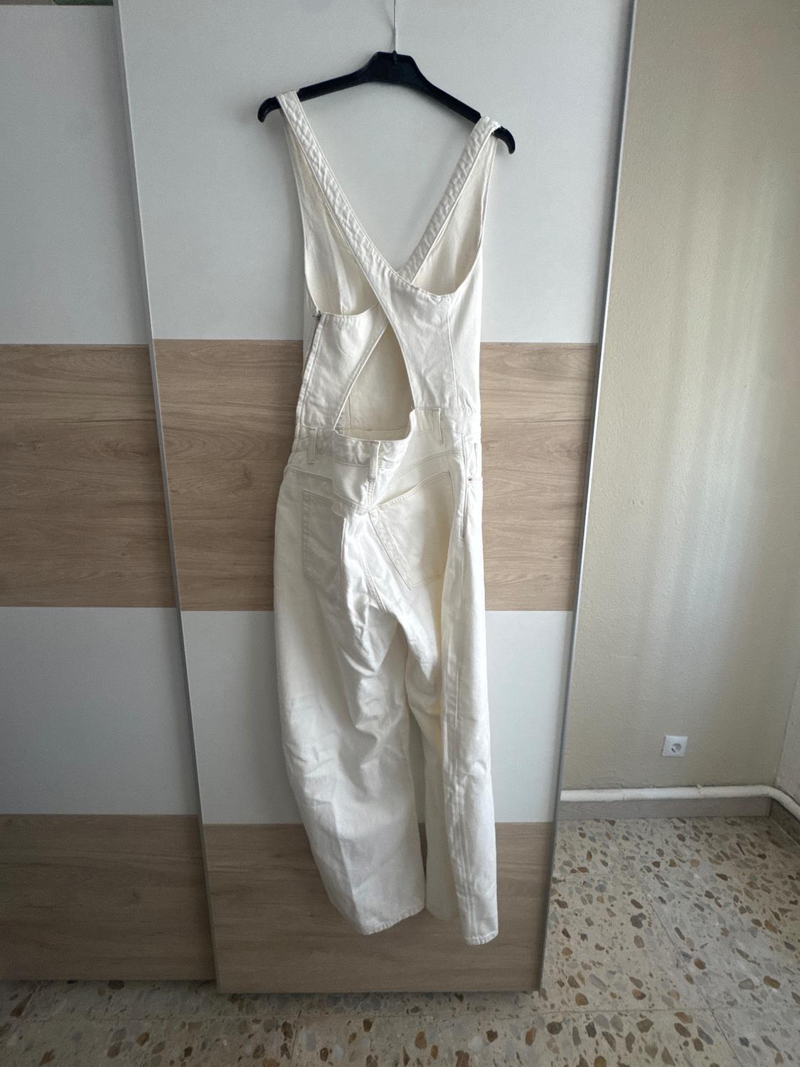 White jumpsuit hanging on a black hanger against a wooden background and white wall. The design features an open back and wide straps.
