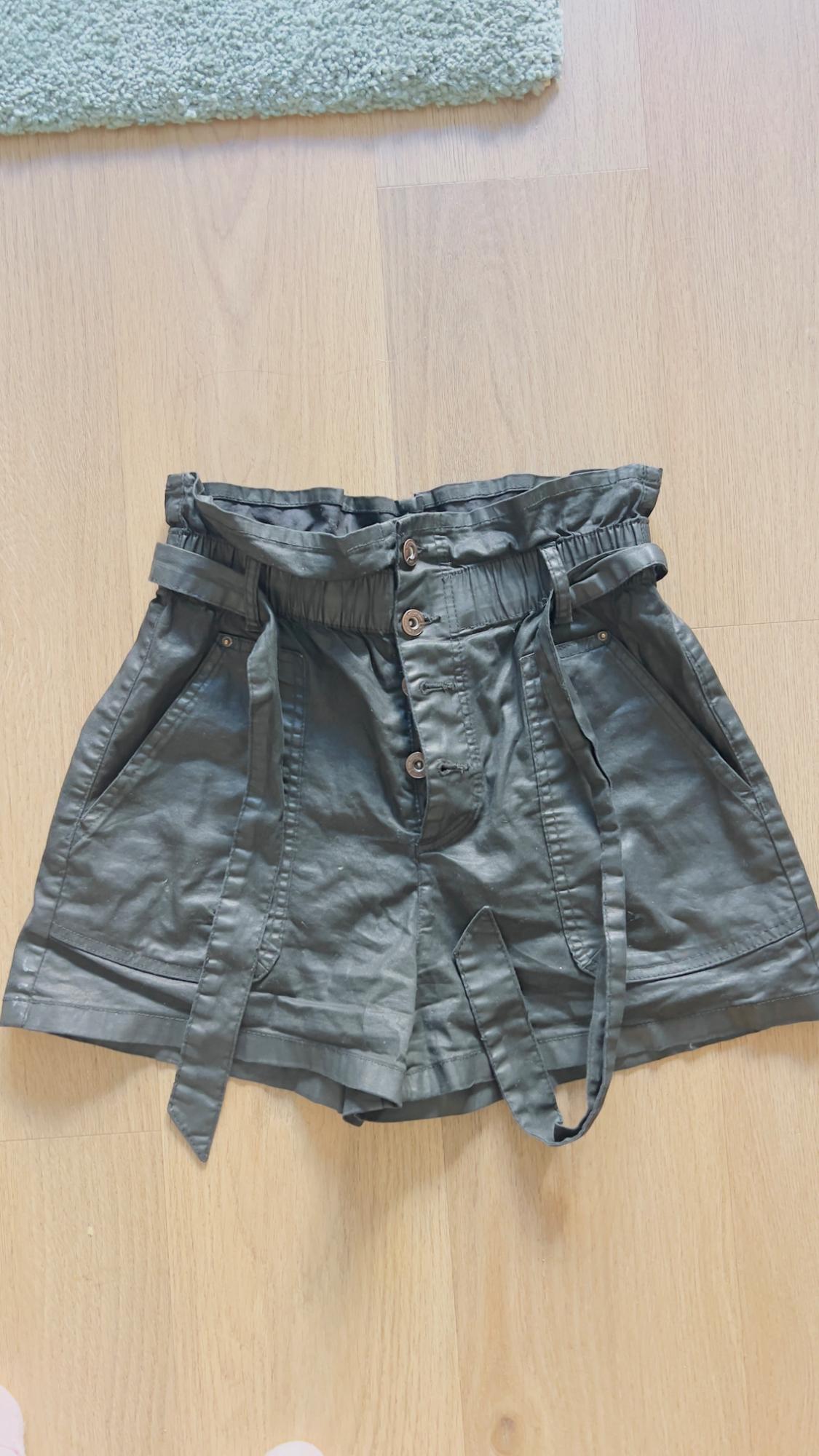 Green shorts with a gathered elastic waistband, button closure, and tie belt, on a light wood floor.