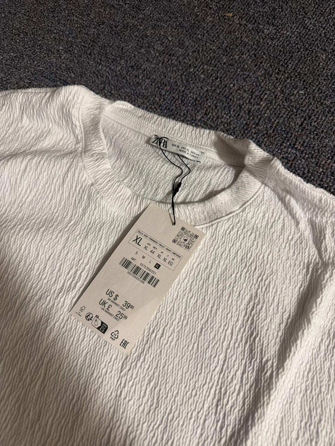 Close-up of the neckline of a textured white t-shirt. Labels with text: ZARA, XL, MADE IN TURKEY, US $ 39.90, UK £ 25.99.
