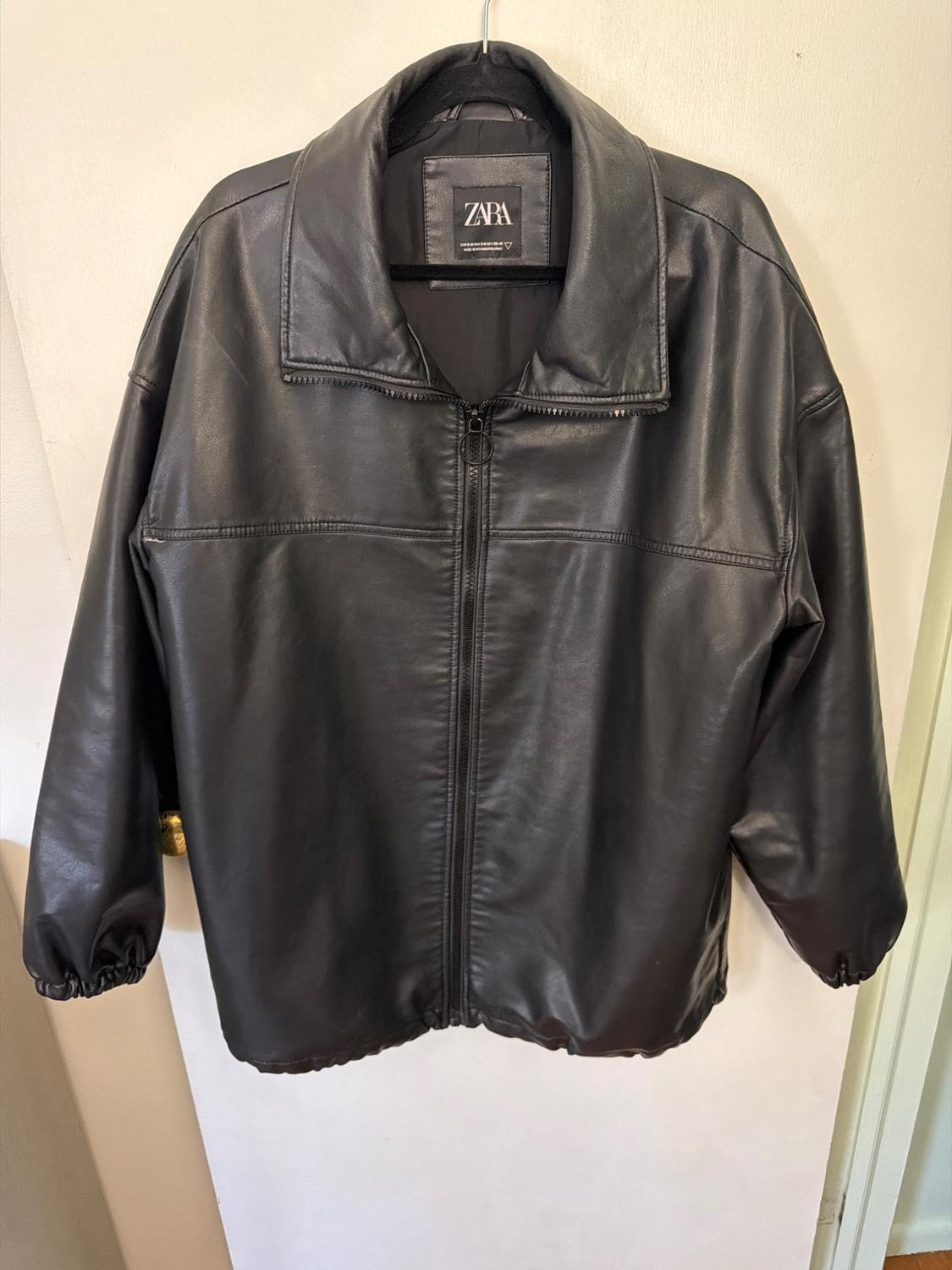 Black jacket with a front zipper and collar, hanging on a hanger over a white door. Interior label with the text ZARA.