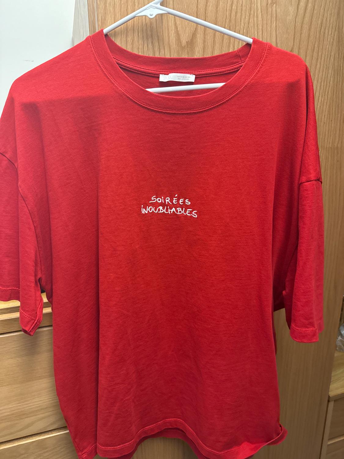 Red t-shirt hanging on a white hanger, with white text on the chest that says "SOIRÉES INOUBLIABLES". Light wood background.