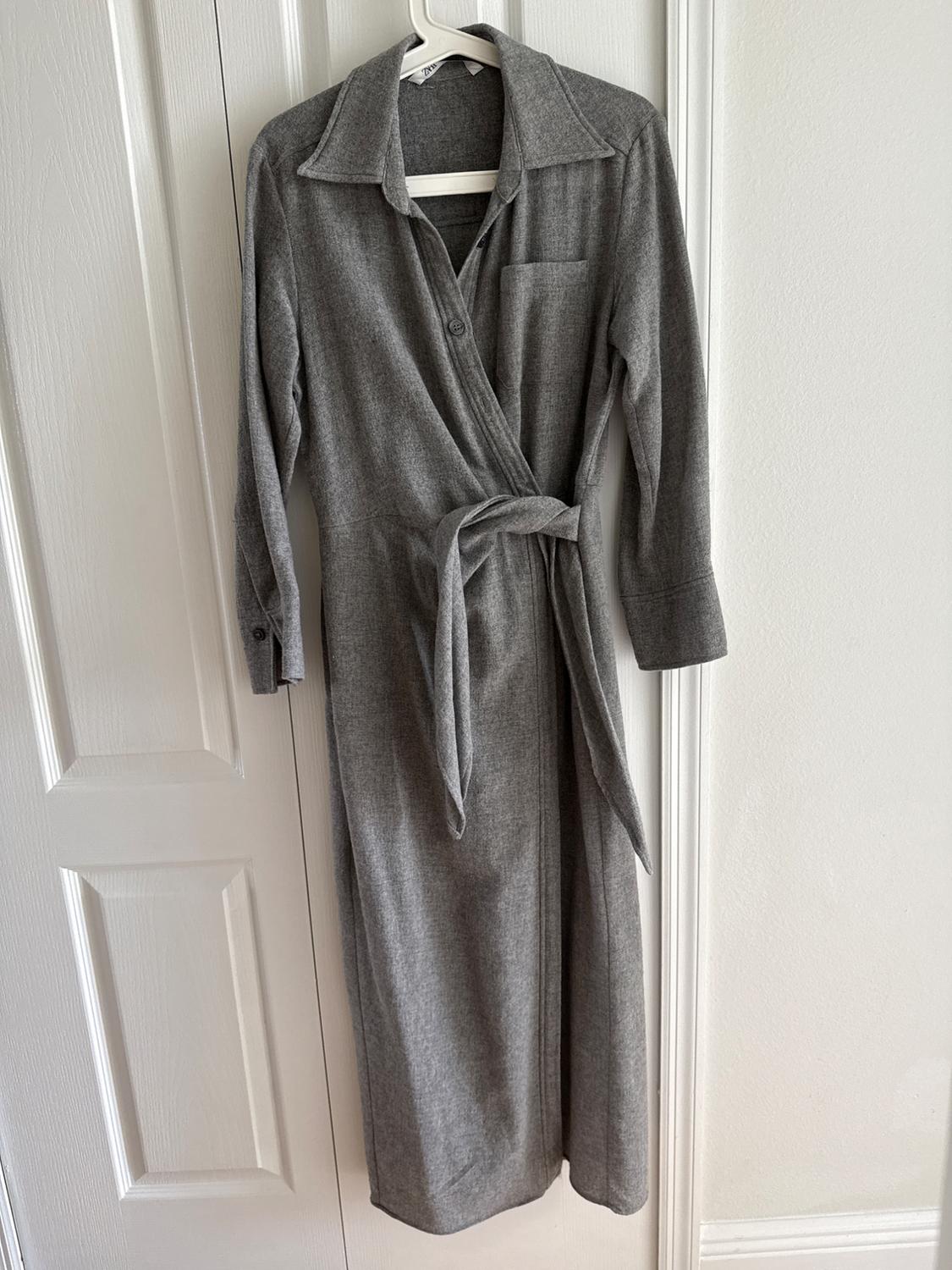Gray long-sleeved dress with a belt, hanging on a white hanger in front of a white door.