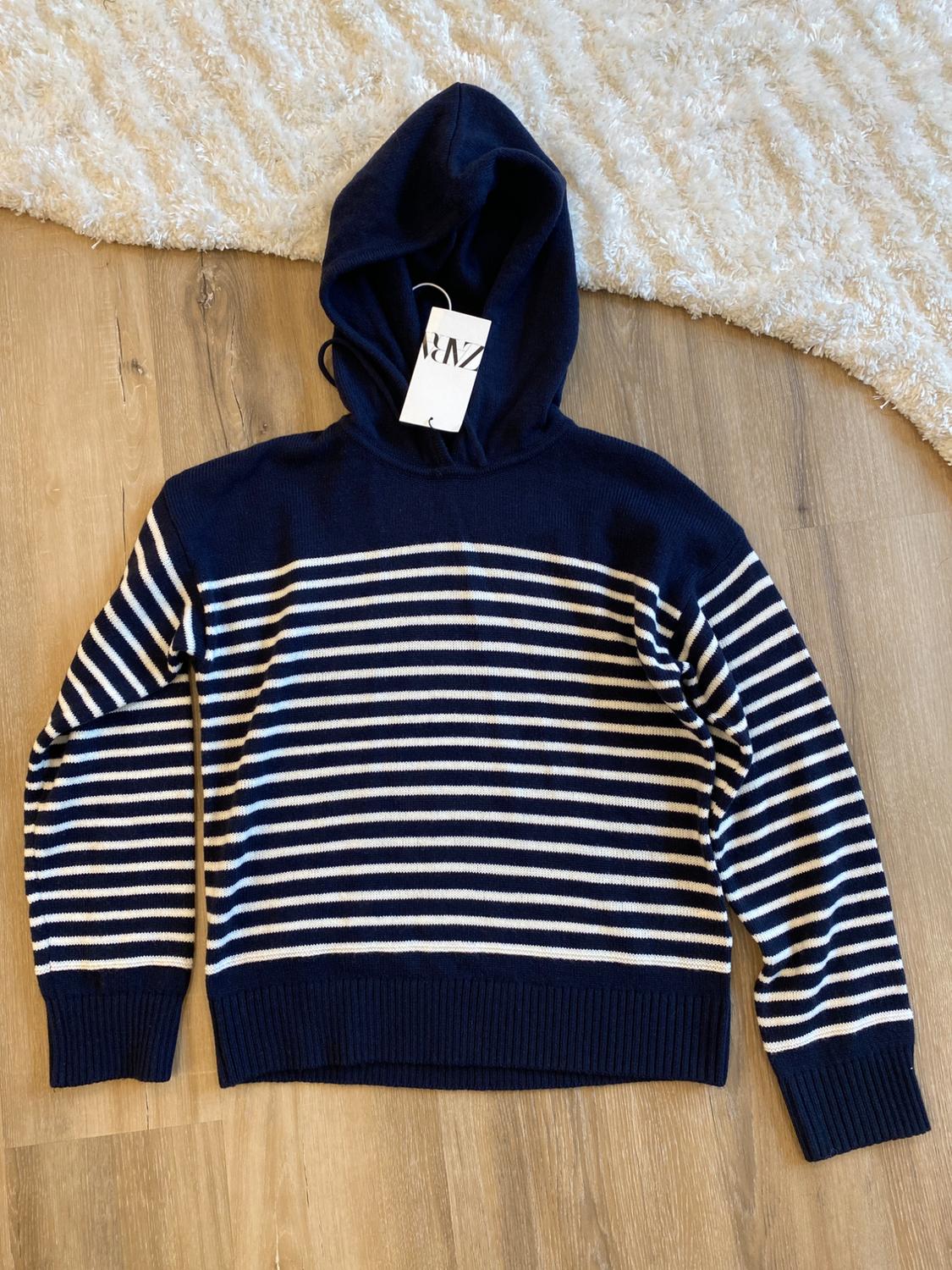 Navy blue hoodie with white stripes on a wooden floor, visible tag hanging from the neck. White rug in the background.