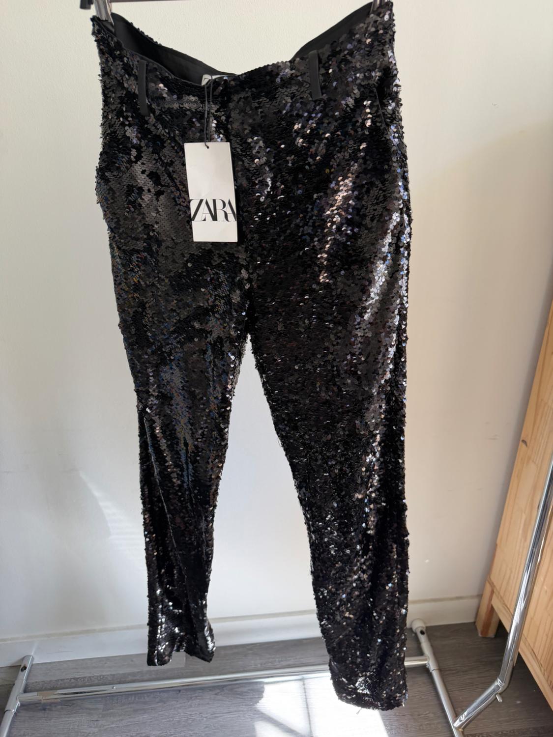 Black sequined trousers hanging on a hanger with a visible Zara tag, against a white background and wooden floor.