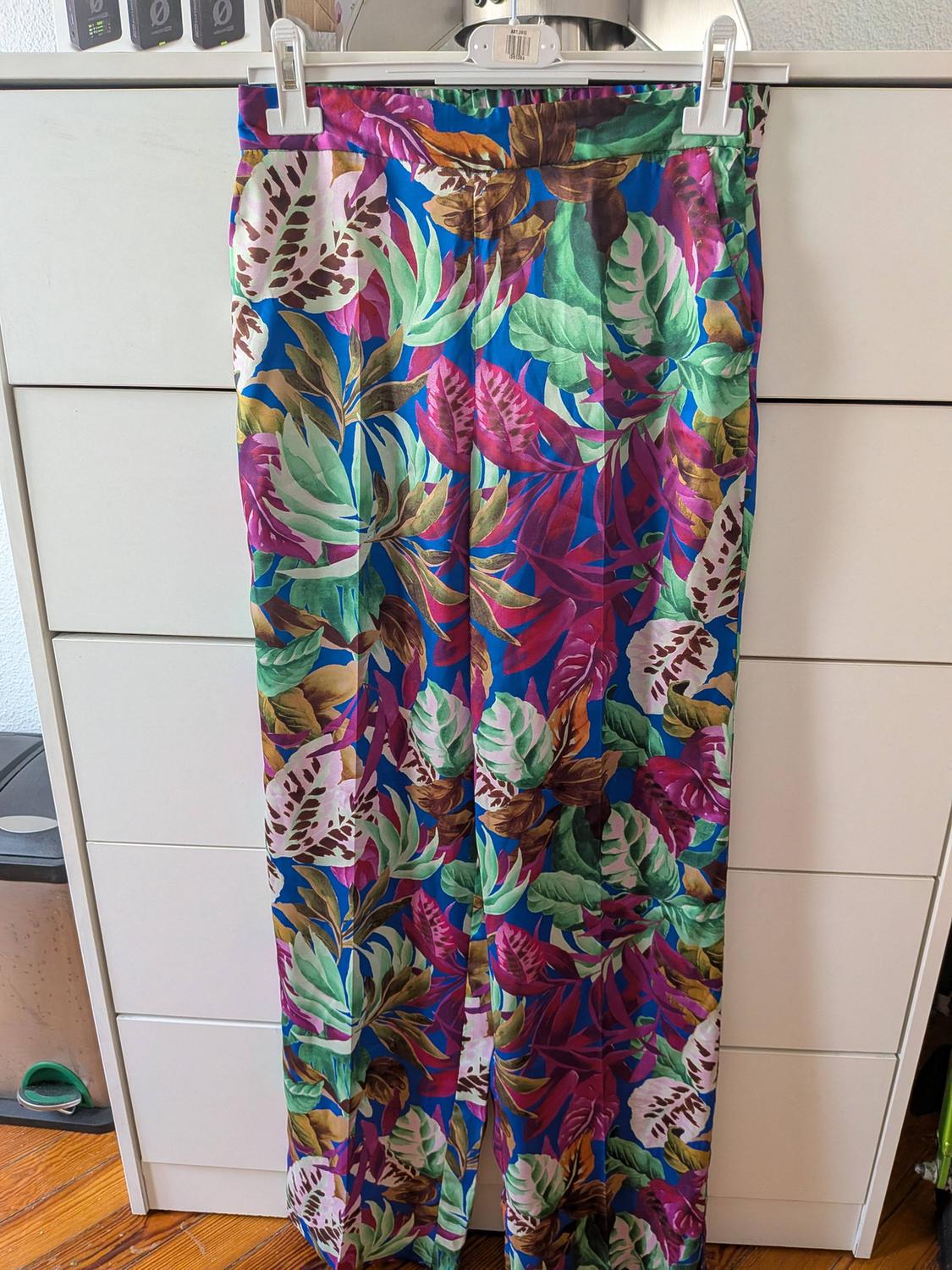 Trousers with a green, pink and brown tropical leaf print on a blue background, hanging on a hanger in front of a white chest of drawers.