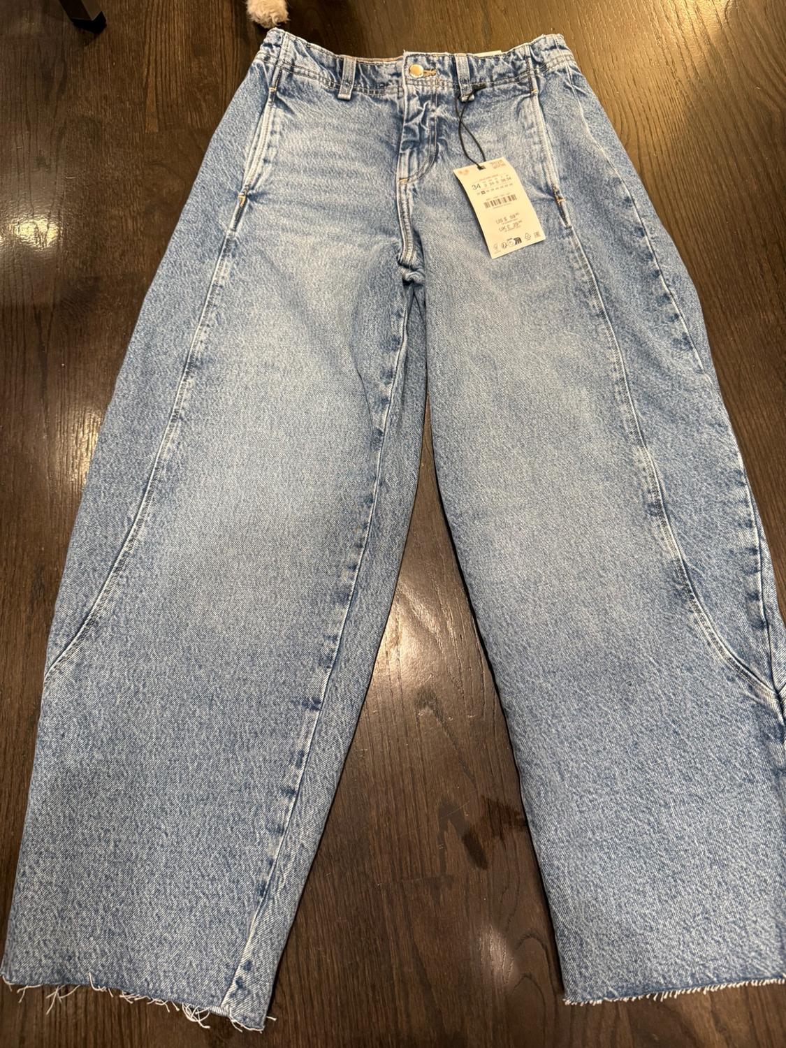 Light-wash, wide-leg denim jeans with front seams and frayed hems on a dark wood floor. Tag with text 34.