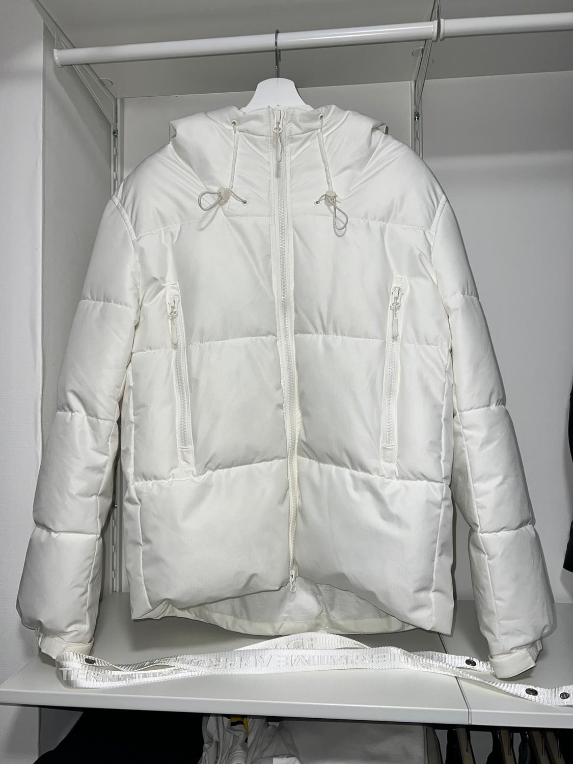 White padded jacket with a hood and zips, hanging on a white hanger inside an open wardrobe.