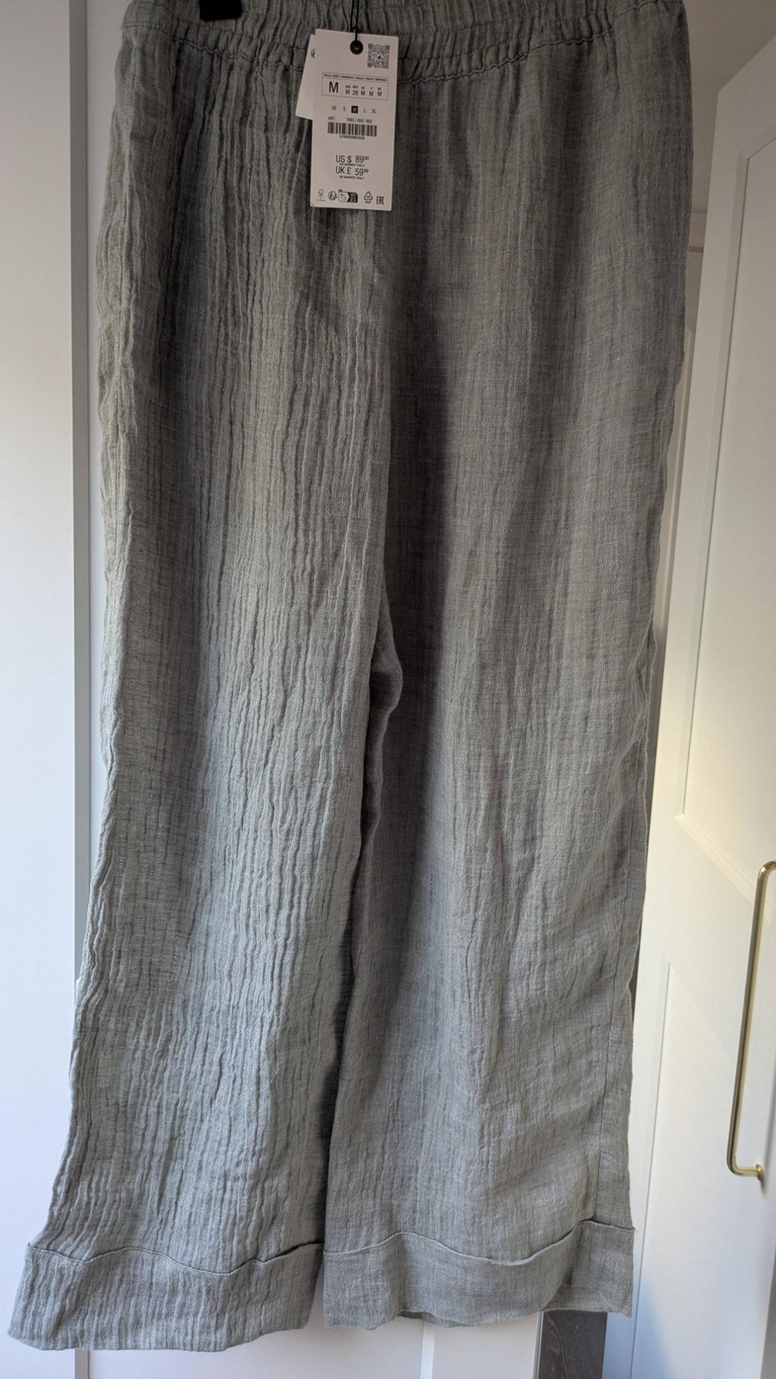 Grey crinkle-fabric trousers hung on a white door, with visible price tag: US $8.99, UK £5.99, size M.