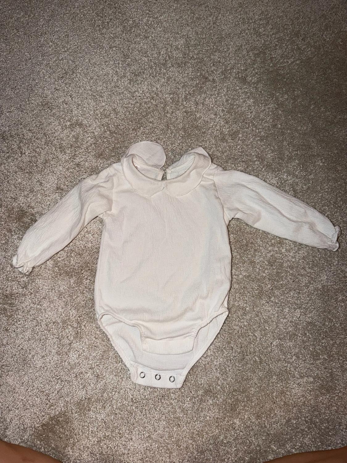 Cream-coloured, long-sleeved baby bodysuit with a rounded collar, laid out on a beige carpet.