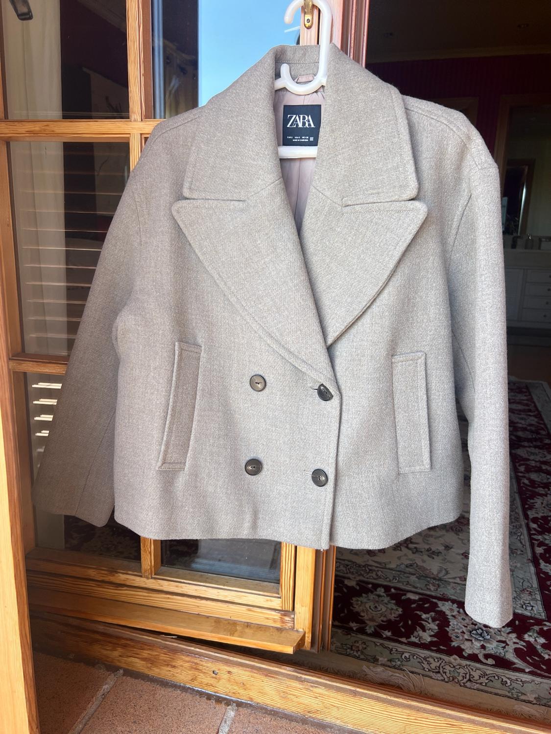 Light grey double-breasted cropped jacket with wide lapels and dark buttons, hanging on a wooden door. ZARA is written on the label.