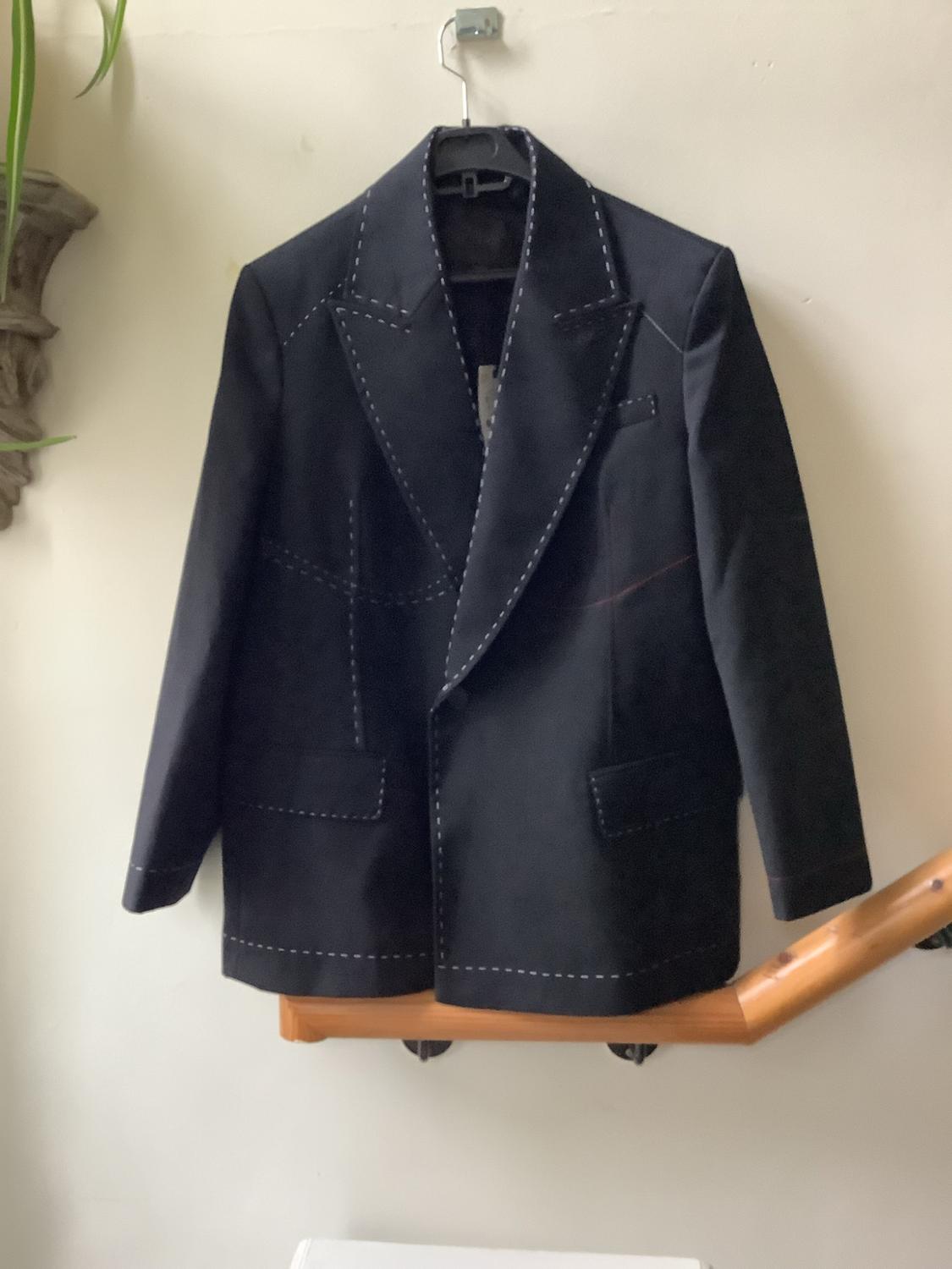 Dark American-style jacket with visible white stitching, hanging on a coat hanger on a white wall above a wooden handrail.