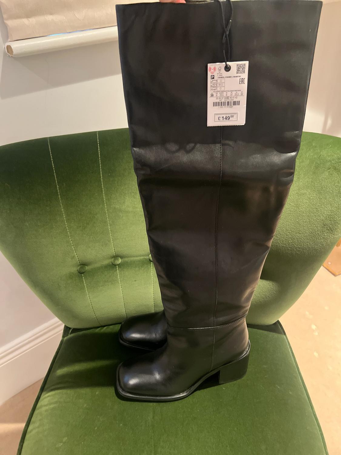 Black knee-high boots with square toes and heels on a green velvet chair. Label with the text “EAC”, “39”, “£149.00”.