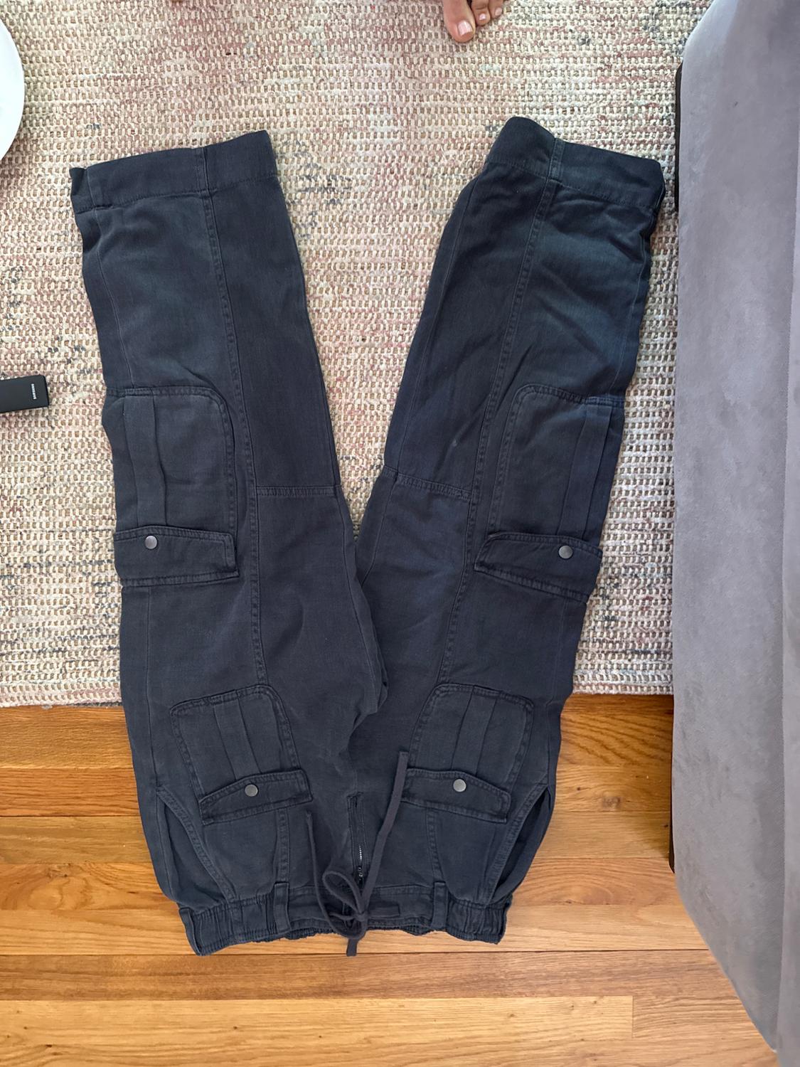 Black cargo pants with large pockets and a drawstring waist, placed on a rug next to a gray sofa.