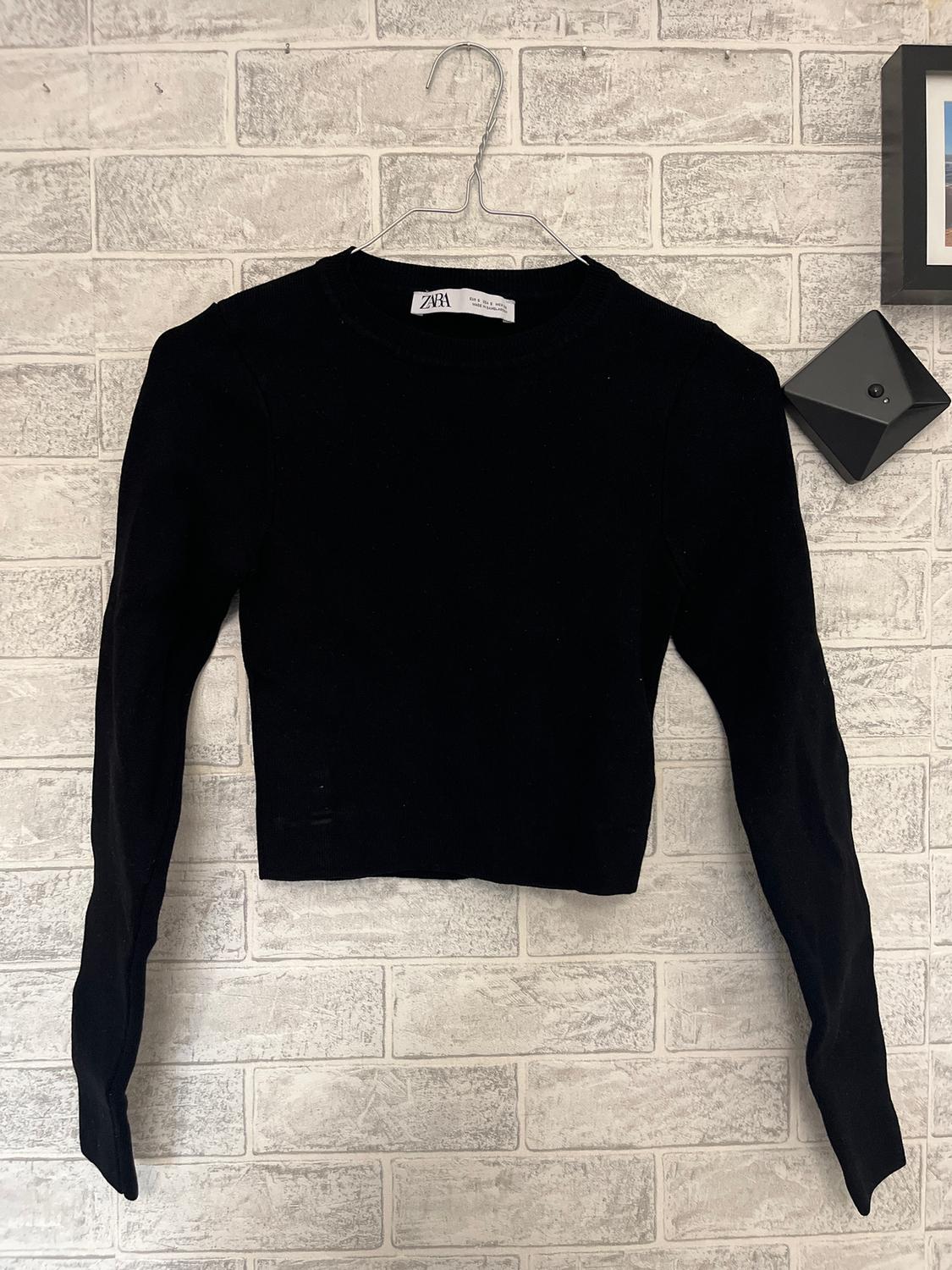 Black long-sleeved jumper hanging on a hanger against a white brick wall. Visible label: "Zara".