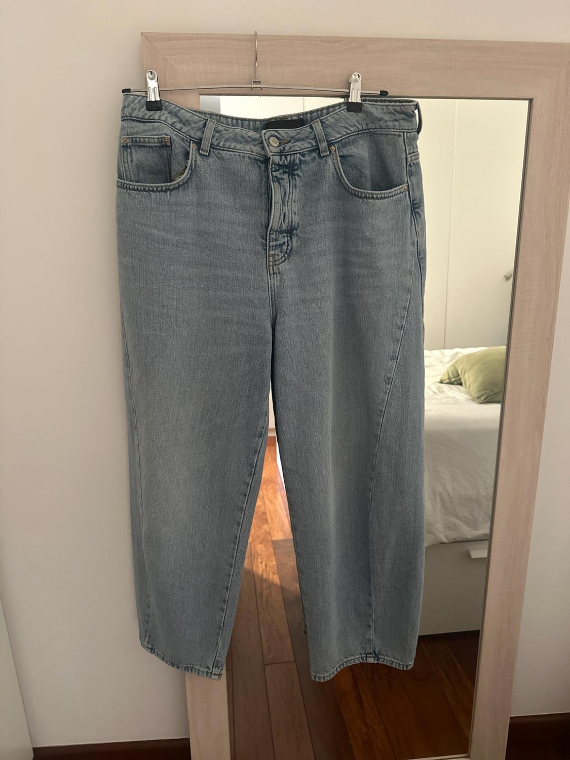 Light blue jeans hanging on a hanger in front of a wooden mirror reflecting a white bed and a wooden floor.