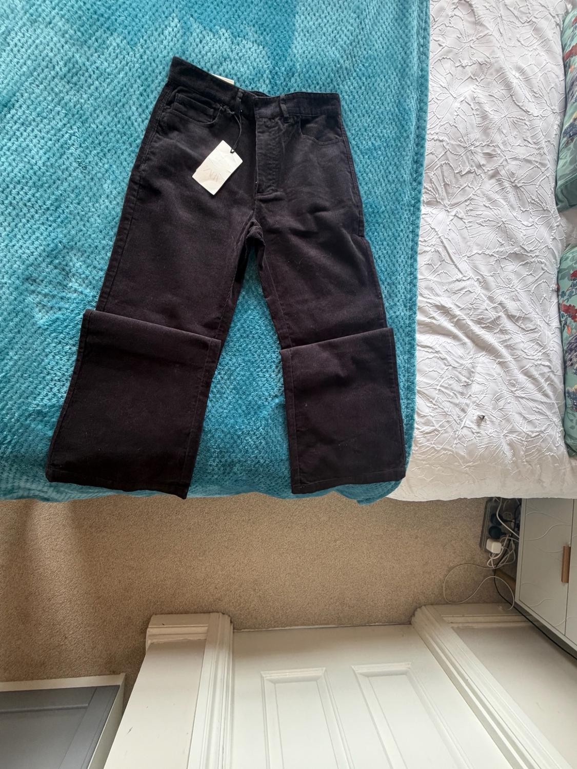 Black trousers with a ZARA label and turned-up hems on a bed with a blue blanket. Casual scene in a bedroom with a beige carpet.