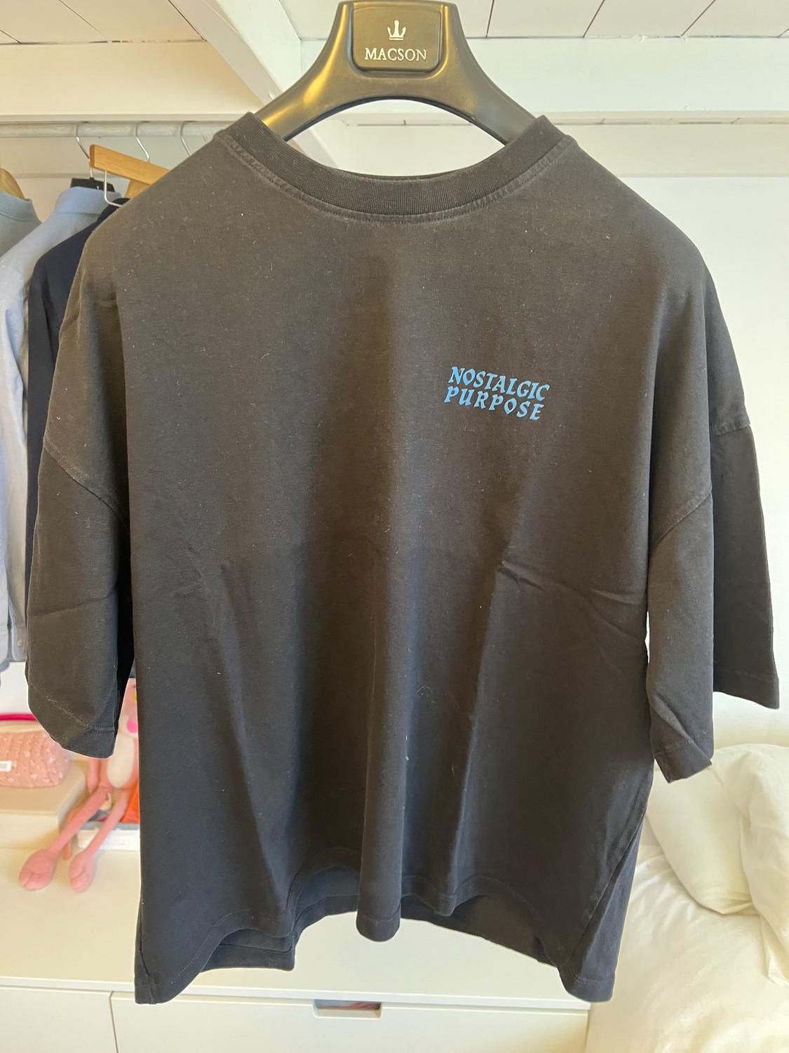 Black T-shirt hanging on a hanger with the text "NOSTALGIC PURPOSE" in blue. Wardrobe background with clothes hanging up.