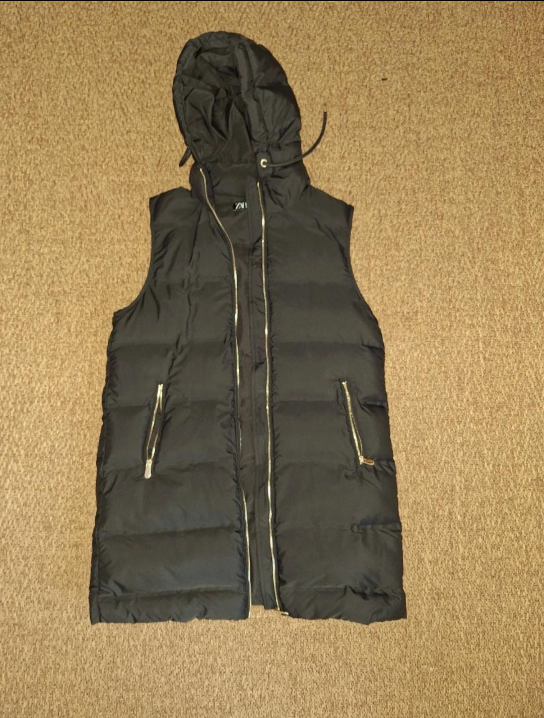 Black padded gilet with hood and zip fastening, on a beige carpet. Pockets with visible zips.