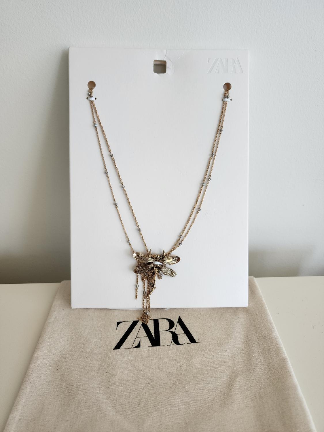 Gold necklace with insect pendant on white cardboard. Background with beige fabric bag with the "ZARA" logo in black.