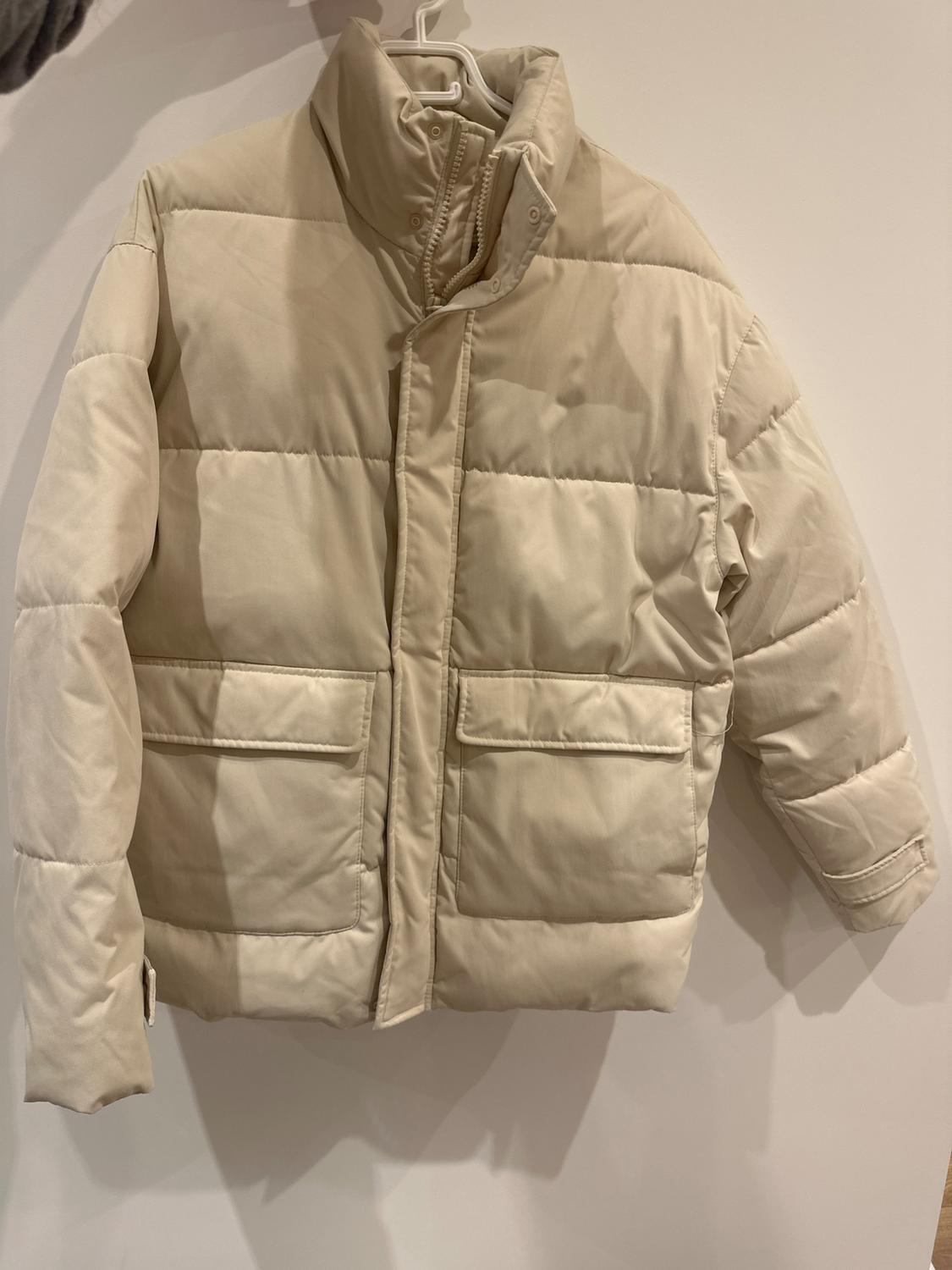 Beige quilted jacket hanging on a white hanger against a light background. Design with a high collar and front pockets.