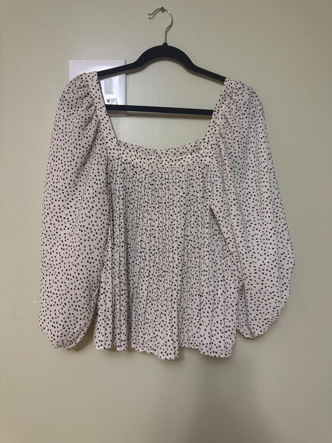 White blouse with black polka dot print, long puffed sleeves, hanging on a black hanger against a beige wall.