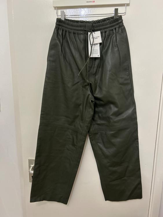 Dark green trousers hanging on a white hanger with a visible tag. White background and door with a metal handle.