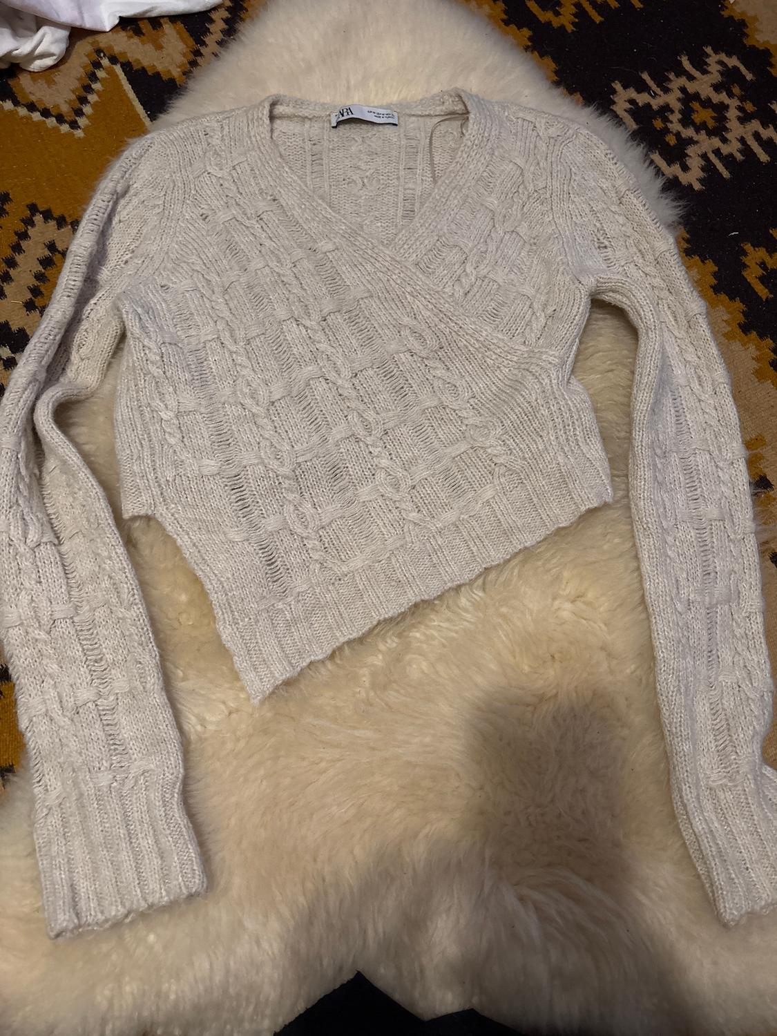 Beige knitted jumper with a cable knit design, long sleeves and a V-neck, on a brown and black geometric patterned rug.