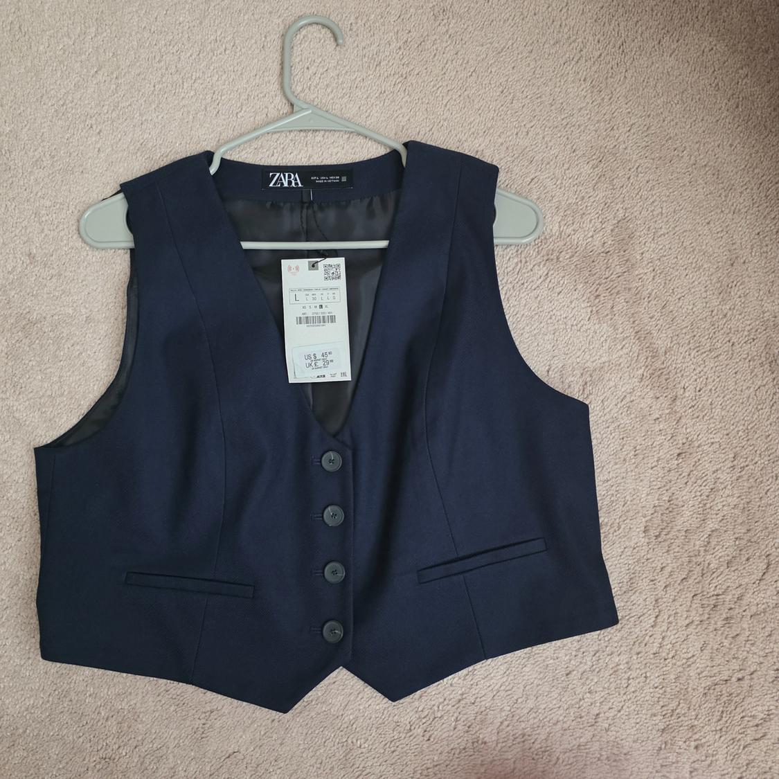 Dark blue Zara vest with front buttons, decorative pockets, and visible tag, hanging on a hanger over a beige carpet.