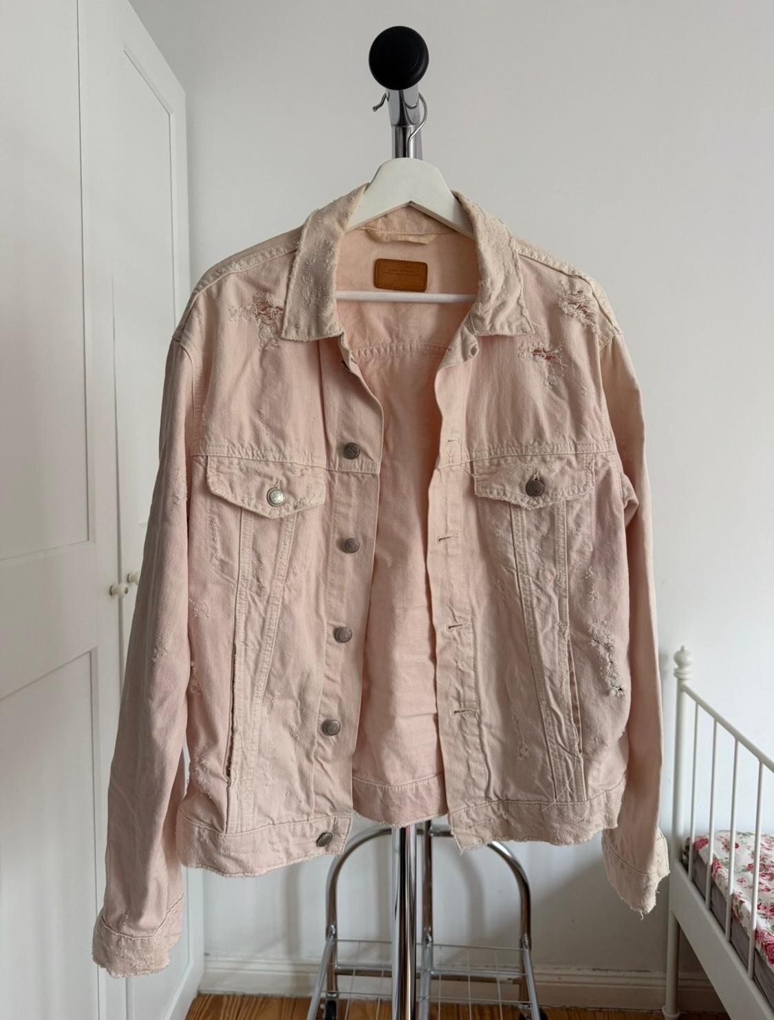 Ανοιχτόχρωμο ροζ denim τζάκετ κρεμασμένο σε λευκή κρεμάστρα, με φθαρμένες λεπτομέρειες, σε ένα δωμάτιο με λευκούς τοίχους και κρεβάτι στο βάθος.