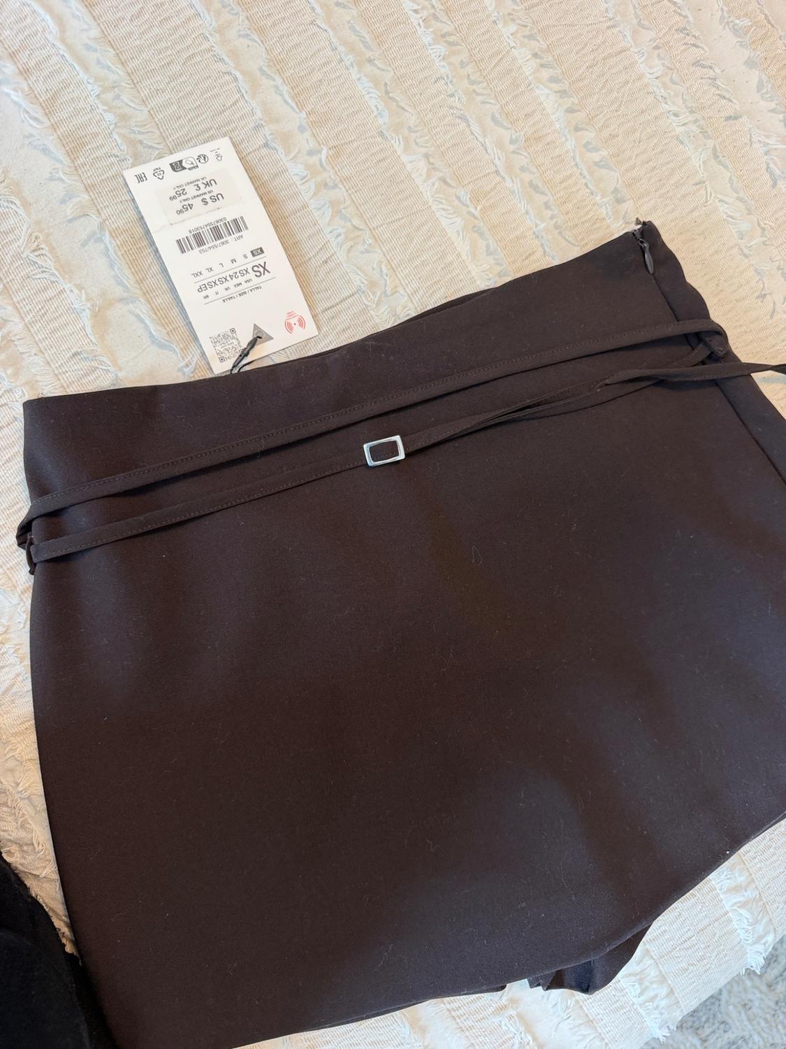 Dark skirt on light-colored fabric with a thin strap and silver buckle. Tag with text: XS ART: 3067/554/753 US $45.90 UK £25.99.