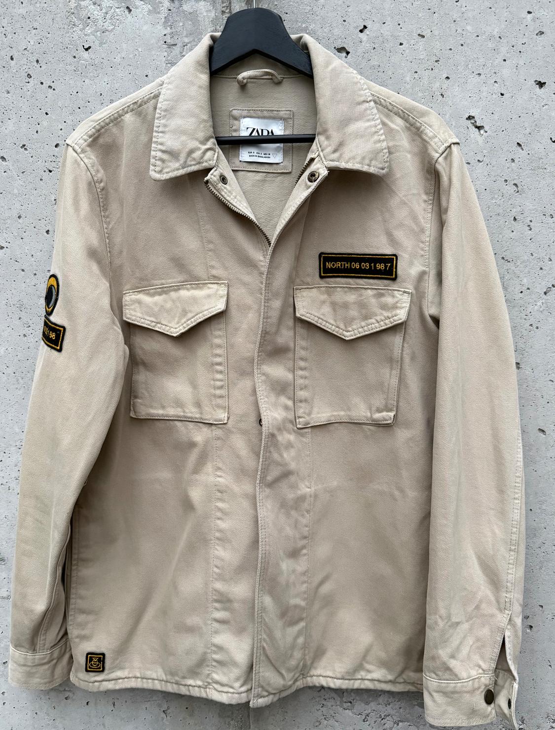 Beige jacket hanging on a black hanger against a concrete background. "NORTH 06 03 1987" patch on the chest and another on the left sleeve.