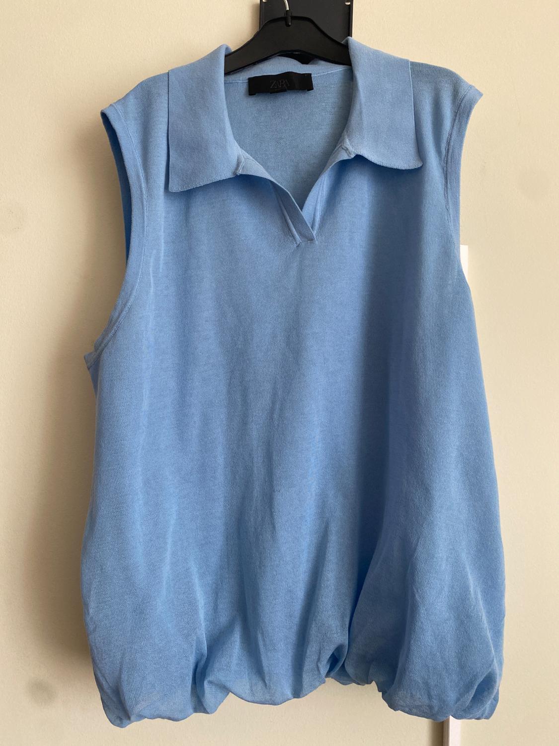 Blue sleeveless gilet with a polo neck, hanging on a black hanger. Visible label with the brand "ZARA". Light beige background.