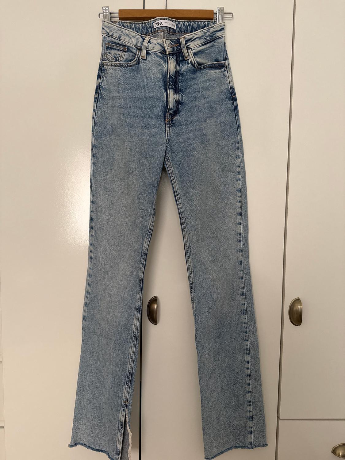 Straight-leg blue jeans hanging on a hanger in front of a white wardrobe. Label visible on the waistband.