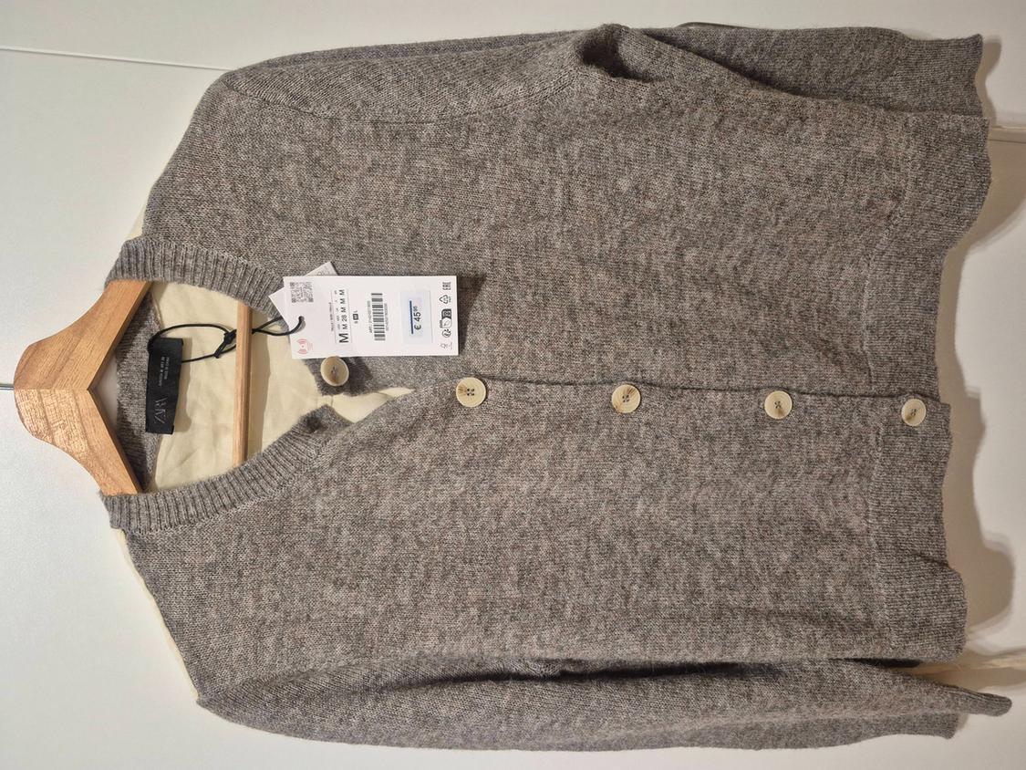 Grey knitted cardigan with buttons on a wooden hanger against a white background. Labels with the text ZARA, M, €45.