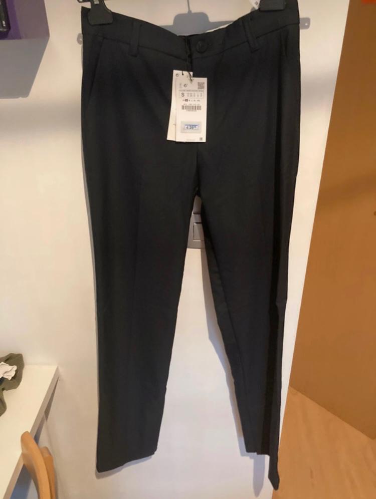 Black trousers hung on a hanger with a visible tag. White wall background and light wooden floor.