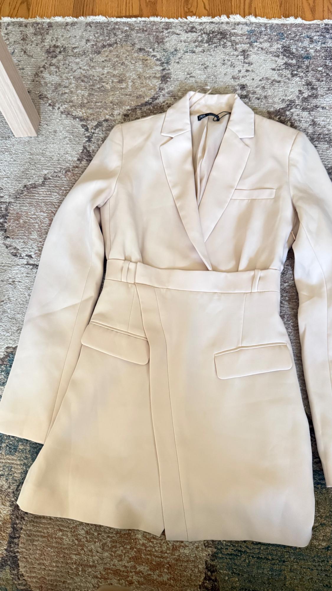 Beige coat with lapels and pockets, placed on a rug with gray and brown tones. Visible tag on the collar.