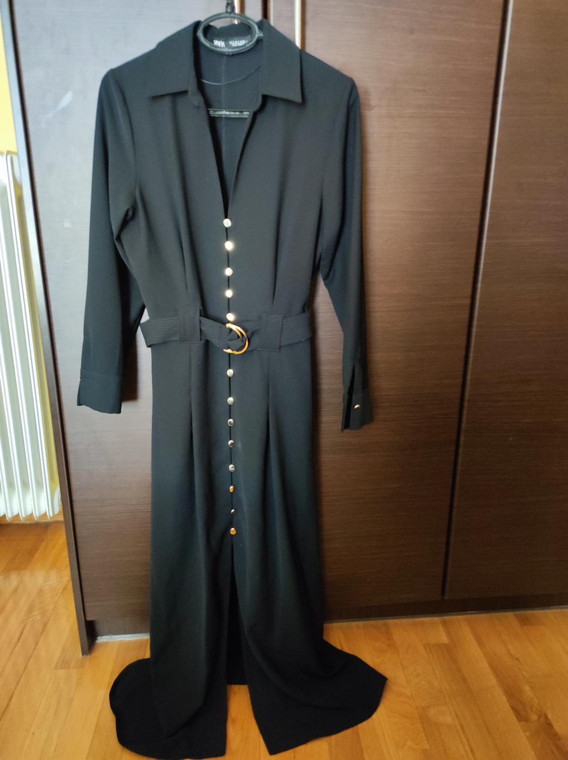 Long black dress with gold buttons and a belt with a gold buckle, hanging on a hanger in front of a wooden wardrobe.