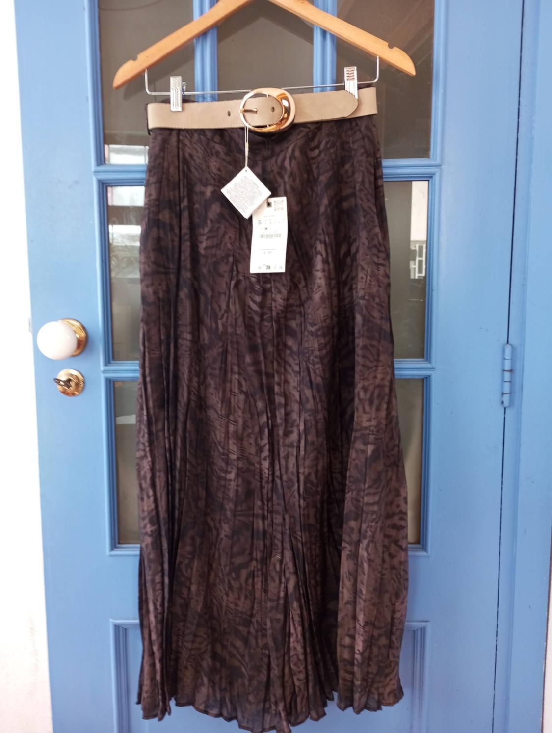 Long brown skirt with abstract print, hanging on a wooden hanger, with a beige belt and visible tags, against a blue door.