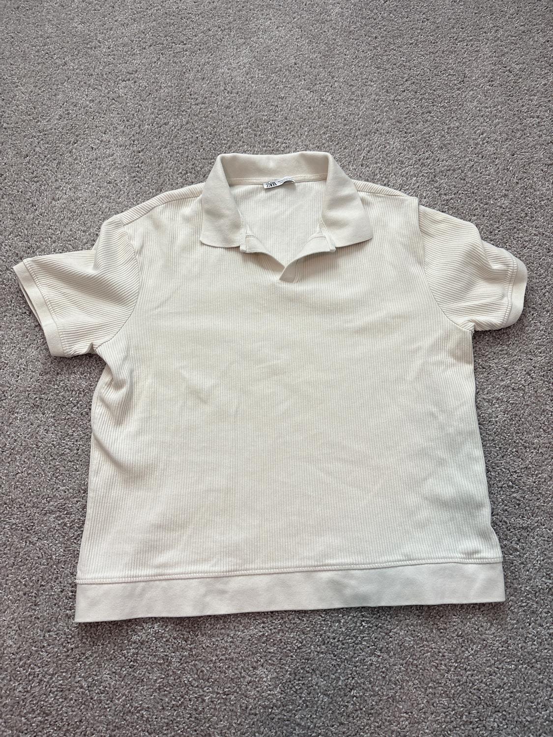 White short-sleeved polo shirt with ribbed texture, placed on a gray carpet. Visible label on the collar.