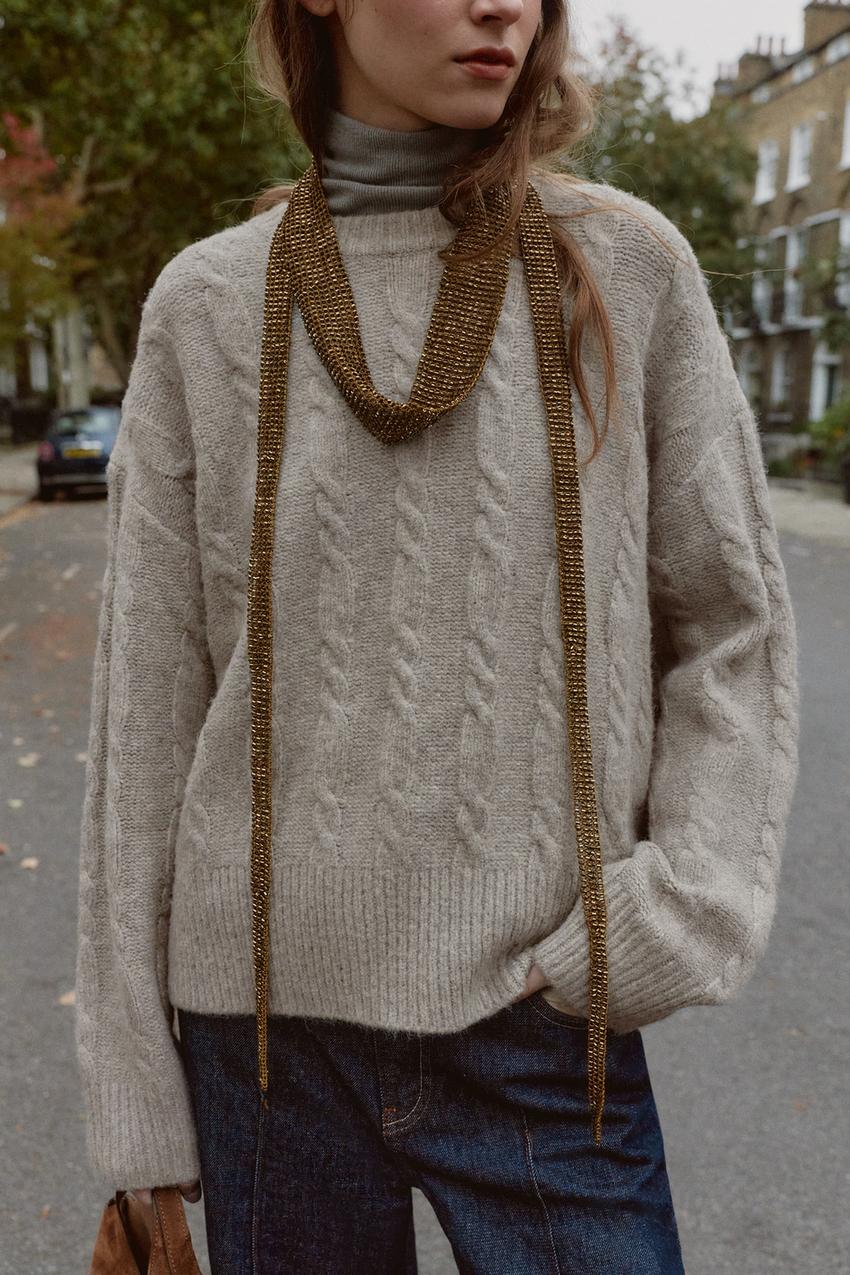 Nahaufnahme eines hellgrauen Strickpullovers mit Rundhalsausschnitt und langen Ärmeln, Zopfmuster, über einem grauen Rollkragenpullover.