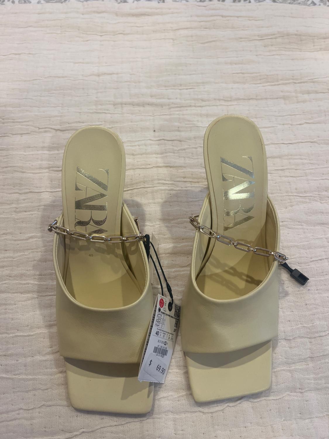 Beige sandals with a square heel, a metal chain and a price tag of $69.90 on wrinkled white fabric.