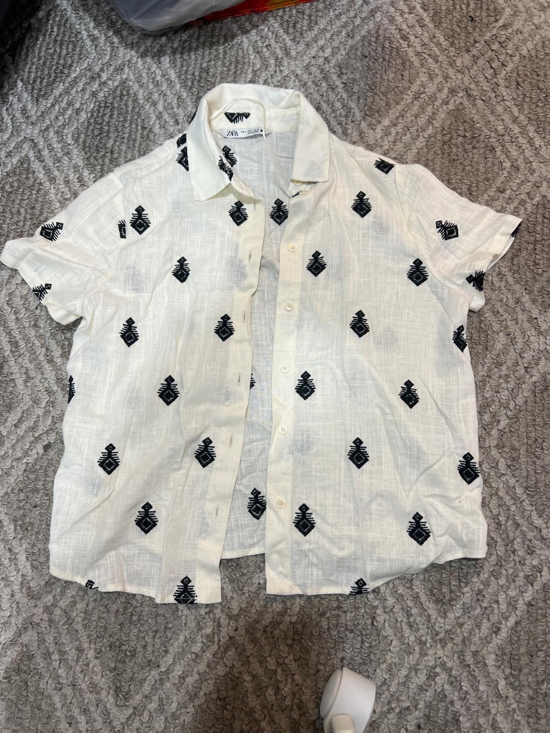 White short-sleeved shirt with black geometric pattern, on a grey carpet. Visible label on the collar.
